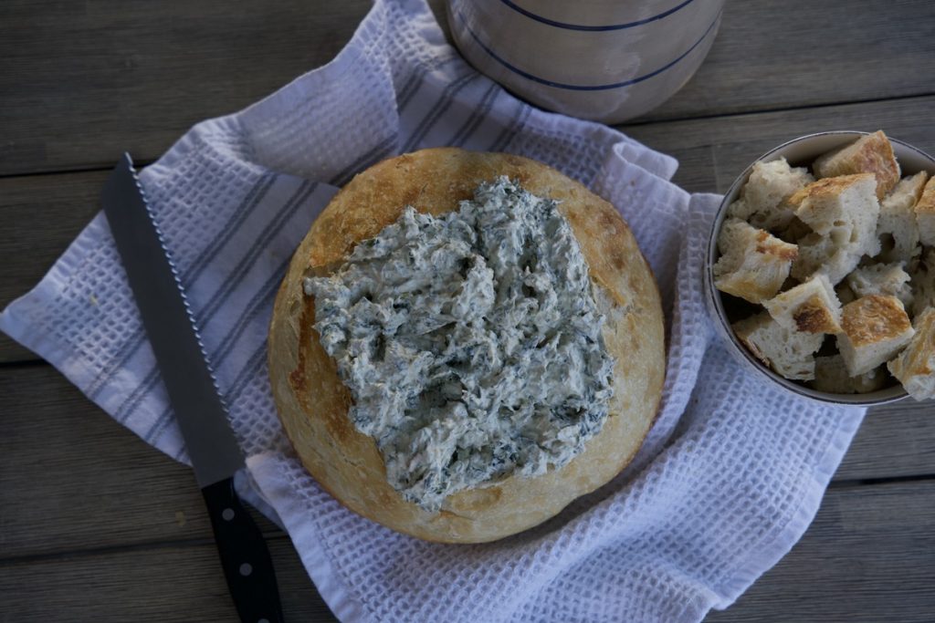 horizontal photo of sourdough bread bowl with spinach dip in the middle on white tea towel. Cut up pieces of sourdough bread beside bread bowl
