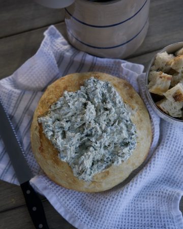 sourdough bread bowl with spinach dip in side on white tea towel. Cut up pieces of sourdough beside bread bowl