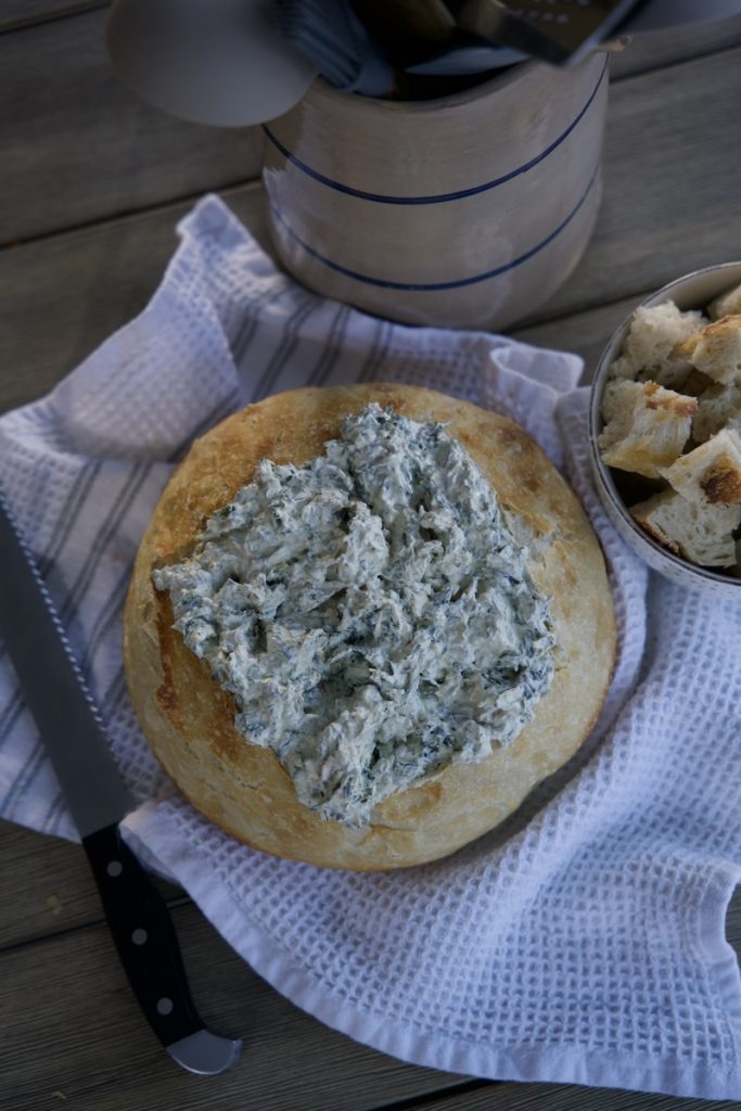sourdough bread bowl with spinach dip in side on white tea towel. Cut up pieces of sourdough beside bread bowl