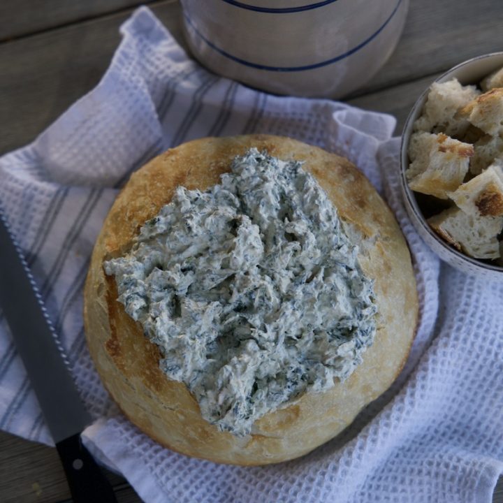 sourdough bread bowl with spinach dip in side on white tea towel. Cut up pieces of sourdough beside bread bowl