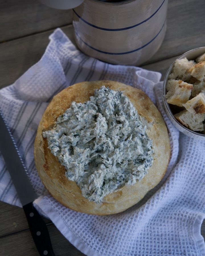sourdough bread bowl with spinach dip in side on white tea towel. Cut up pieces of sourdough beside bread bowl
