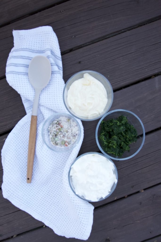 spinach dip ingredients in small bowls in wood table with white tea towel and large wood spoon beside it
