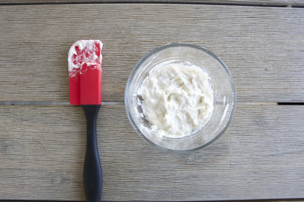 A small glass bowl with 1 heaping tablespoon of all purpose flour and 1 tablespoon of water mixed together with red icing spatula beside it, sitting on wooden table.