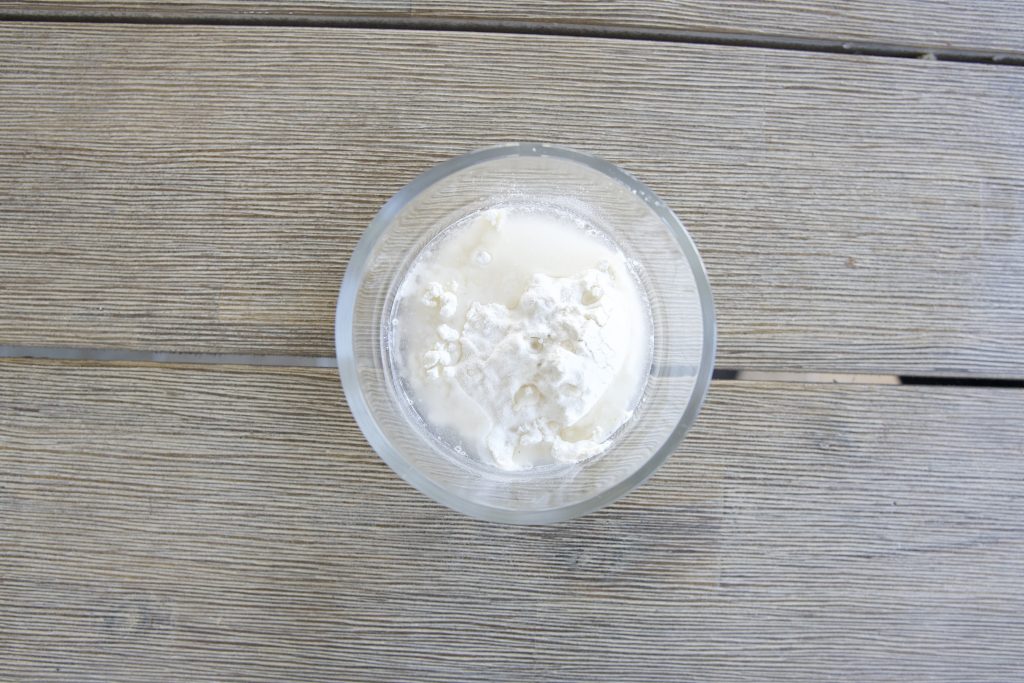 small glass bowl on wooden table with 1 heaping tablespoon flour and 1 tablespoon water on top, not mixed for simple sourdough starter recipe.