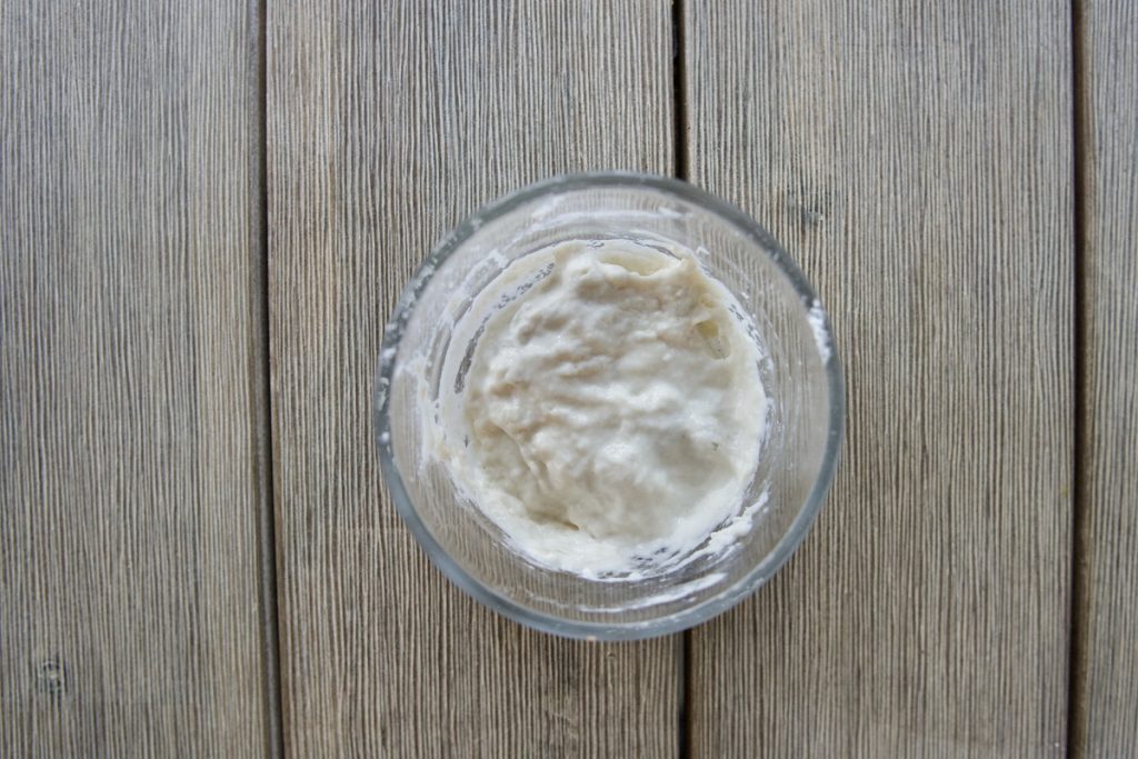 small glass bowl on wooden table with 1 heaping tablespoon flour and 1 tablespoon water mixed together on day 2 for simple sourdough starter recipe