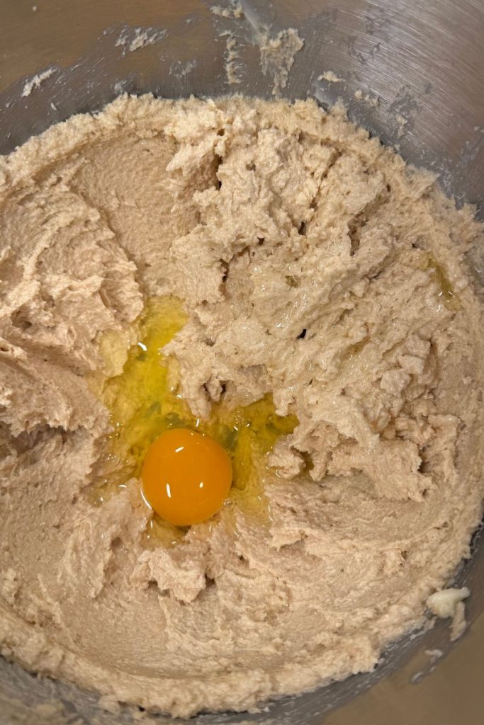 egg added to butter and sugar and vanilla in a silver kitchen aid mixer bowl for oatmeal chocolate chip cookie dough recipe